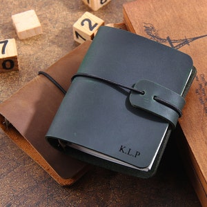 Puede incluir: Se muestran dos diarios de cuero, uno marrón y otro verde oscuro. El diario verde tiene las iniciales "K.L.P." grabadas en la portada. Bloques de madera con números y una caja de madera con texto y una ilustración de la Torre Eiffel están en el fondo.