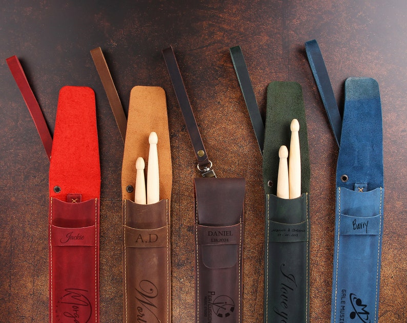 Peut inclure: Cinq porte-baguettes en cuir personnalis&eacute;s de diff&eacute;rentes couleurs et avec des noms. Les porte-baguettes sont en cuir marron, rouge, vert, bleu et noir. Chaque porte-baguettes a une sangle et une poche pour les baguettes. Les noms sur les porte-baguettes sont Jackie, A.D., Daniel, Je t'aime et Barry.