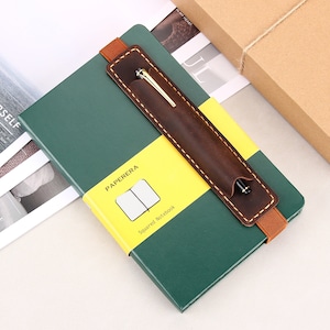 May include: A green leather-bound notebook with a brown leather pen holder and a yellow label that says "PAPERERA Squared Notebook".