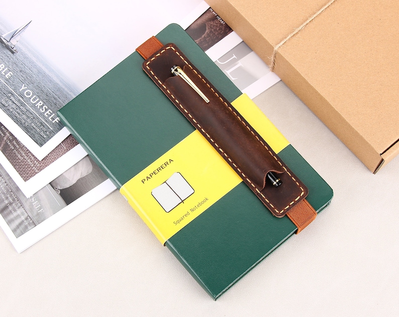 May include: A green leather-bound notebook with a brown leather pen holder and a yellow label that says "PAPERERA Squared Notebook".