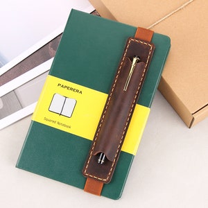 May include: A green leather-bound notebook with a yellow label that says "PAPERERA Squared notebook". A brown leather pen holder with a gold pen is attached to the notebook.