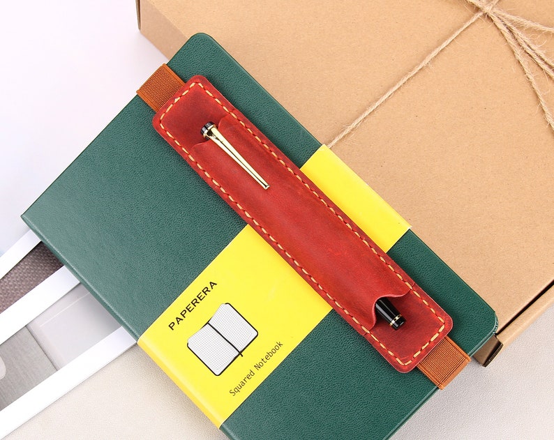 May include: A green notebook with a yellow and red leather pen holder. The notebook has the text "PAPERERA" and "Squared Notebook" printed on it.
