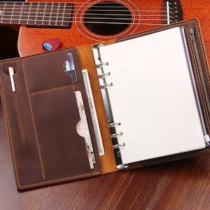 May include: A brown leather bound journal with a zippered pocket and a pen holder. The journal has a ring binder system and lined pages.