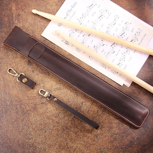 Peut inclure: Un &eacute;tui &agrave; baguettes en cuir marron avec une sangle et un porte-cl&eacute;s. L'&eacute;tui est pos&eacute; sur une surface en bois avec deux baguettes en bois et une partition de musique.