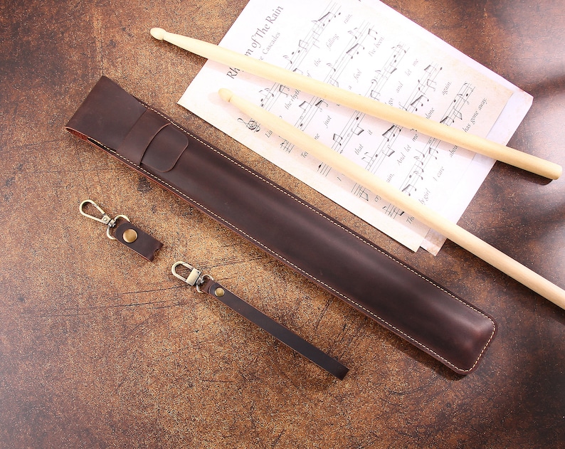 Peut inclure: Un &eacute;tui &agrave; baguettes en cuir marron avec une sangle et un porte-cl&eacute;s. L'&eacute;tui est pos&eacute; sur une surface en bois avec deux baguettes en bois et une partition de musique.