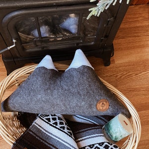 May include: A grey mountain-shaped cushion with white peaks and a brown leather patch featuring a tree and the text "Wilderness". The cushion is in a wicker basket with a black and white striped blanket and a green candle with the word "Wild" on it.