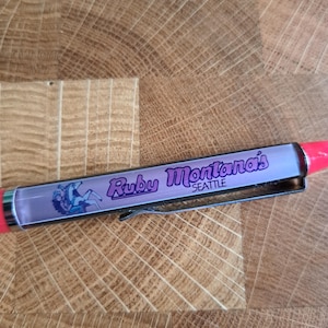Può includere: Una penna nera e rossa con corpo trasparente. La penna presenta una grafica di una donna a cavallo con il testo "Ruby Montana's Seattle" stampato sul lato.
