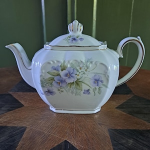 Könnte beinhalten: Weiße Porzellan-Teekanne mit floralem Design mit lila Blumen und grünen Blättern. Die Teekanne hat einen goldenen Rand und einen passenden Deckel. Der Griff und der Ausguss sind gebogen. Ein klassischer Artikel für das Teeservice.