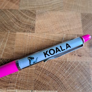 Könnte beinhalten: Ein rosa Stift mit einem silbernen Band, auf dem ein Koala abgebildet ist und das Wort "KOALA" aufgedruckt ist.