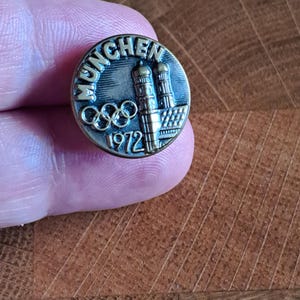 Könnte beinhalten: Eine Vintage-Anstecknadel aus Bronze mit dem Wort "MUNCHEN" über den olympischen Ringen und dem Jahr "1972". Die Anstecknadel zeigt zwei stilisierte Türme und ein detailliertes Design, wahrscheinlich zur Erinnerung an die Olympischen Spiele 1972 in München.