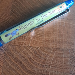 Può includere: Una penna blu con un'etichetta gialla e arancione che dice "Runnin' in the Sun ARIZONA". L'etichetta presenta anche un roadrunner dei cartoni animati.