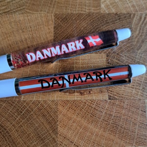 Könnte beinhalten: Zwei weiße Stifte mit rotem Glitzer und dem Wort "DANMARK" in weiß auf rotem Hintergrund. Die Stifte zeigen auch die dänische Flagge.