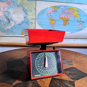 May include: A red kitchen scale with a black face and a green dial. The scale has a maximum weight capacity of 5 kg or 11 lbs. The scale is sitting on a wooden table with a starburst pattern.