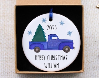 Pode incluir: Enfeite de Natal em cerâmica branca com um camião azul a transportar uma árvore de Natal verde. O enfeite tem o ano de 2025 e o texto "Merry Christmas William" com flocos de neve azuis. Apresentado numa caixa de presente castanha.