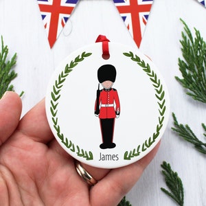Könnte beinhalten: Weißer Keramik-Weihnachtsbaumschmuck mit einem grünen Kranzdesign. Der Anhänger zeigt eine Comic-Illustration eines britischen Wachmanns in roter Uniform und einem schwarzen Bärenfellhut. Der Text "James" ist unter der Illustration gedruckt.