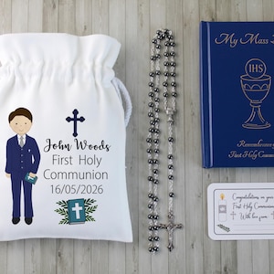 K&ouml;nnte beinhalten: Ein Erstkommunion-Set mit einem wei&szlig;en Kordelzugbeutel mit einer Jungenillustration und Text, einem Rosenkranz, einem blauen Buch mit goldener Schrift und einer Karte. Der Beutel tr&auml;gt den Namen "John Woods" und das Datum 16.05.2026.
