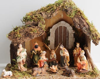 May include: A nativity scene set featuring a wooden stable structure with a bark and moss roof. Inside are hand-painted figurines of the Holy Family, an angel, three kings, a shepherd, and animals including a sheep, a donkey, and a cow.