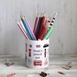Könnte beinhalten: Weißer Stifthalter mit London-Motiven und dem Text "Noah's pencil pot". Der Behälter ist mit farbigen Stiften gefüllt. Mehrere Büroklammern und eine rote Büroklammer liegen auf der Holzoberfläche.