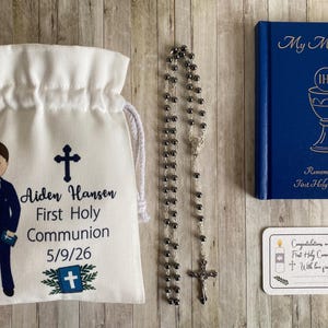K&ouml;nnte beinhalten: Ein Erstkommunion-Set mit einem wei&szlig;en Kordelzugbeutel mit einer Jungenillustration und Text, einem Rosenkranz, einem blauen Buch mit dem Titel "My Mass Book" und einer Karte. Der Beutel tr&auml;gt den Namen "Aiden Hansen" und das Datum.