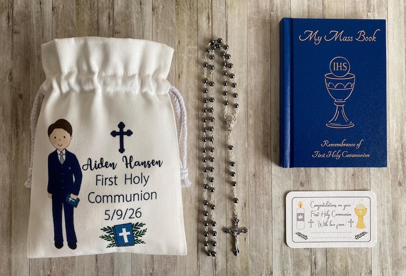 K&ouml;nnte beinhalten: Ein Erstkommunion-Set mit einem wei&szlig;en Kordelzugbeutel mit einer Jungenillustration und Text, einem Rosenkranz, einem blauen Buch mit dem Titel "My Mass Book" und einer Karte. Der Beutel tr&auml;gt den Namen "Aiden Hansen" und das Datum.