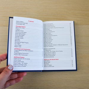 K&ouml;nnte beinhalten: Ein offenes Buch mit blauem Einband, das ein Inhaltsverzeichnis mit verschiedenen Gebetstiteln in roter und schwarzer Schrift zeigt. Das Buch wird von einer Hand mit hellem Nagellack offen gehalten. Der Hintergrund ist eine hellbraune Holzoberfl&auml;che.