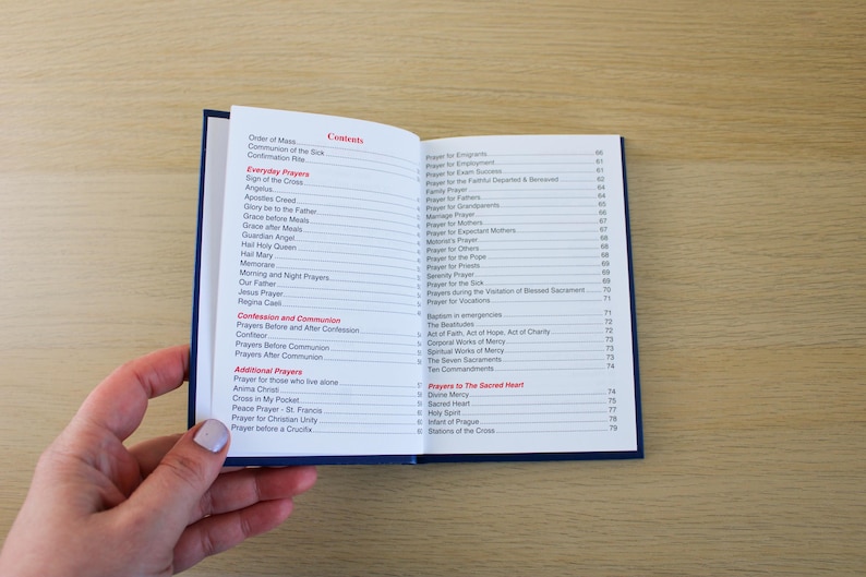 K&ouml;nnte beinhalten: Ein offenes Buch mit blauem Einband, das ein Inhaltsverzeichnis mit verschiedenen Gebetstiteln in roter und schwarzer Schrift zeigt. Das Buch wird von einer Hand mit hellem Nagellack offen gehalten. Der Hintergrund ist eine hellbraune Holzoberfl&auml;che.