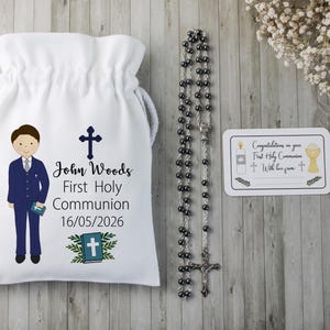 K&ouml;nnte beinhalten: Ein wei&szlig;er Beutel mit Kordelzug, der eine Cartoon-Illustration eines Jungen in einem blauen Anzug, ein Kreuz und den Text "John Woods Erste Heilige Kommunion 16/05/2026" zeigt. Ein Rosenkranz und eine Karte mit religi&ouml;sen Symbolen sind ebenfalls vorhanden.