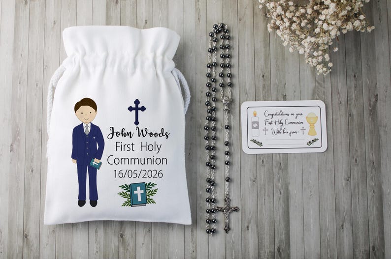 K&ouml;nnte beinhalten: Ein wei&szlig;er Beutel mit Kordelzug, der eine Cartoon-Illustration eines Jungen in einem blauen Anzug, ein Kreuz und den Text "John Woods Erste Heilige Kommunion 16/05/2026" zeigt. Ein Rosenkranz und eine Karte mit religi&ouml;sen Symbolen sind ebenfalls vorhanden.
