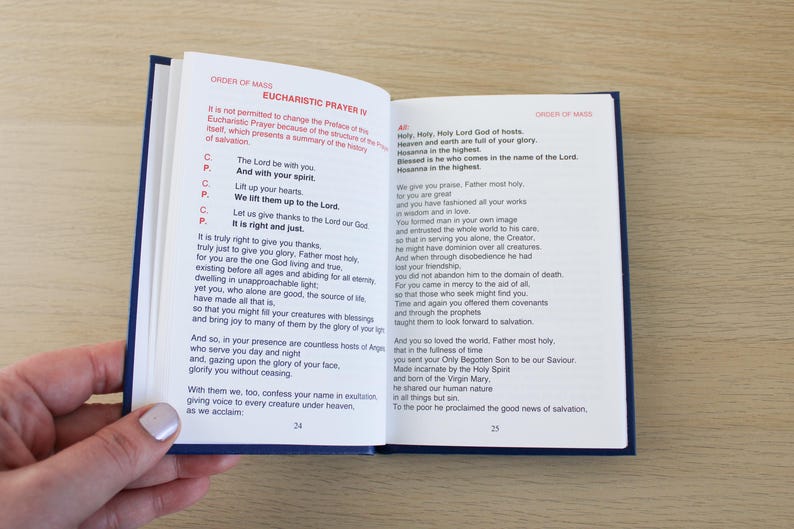 K&ouml;nnte beinhalten: Offenes Buch mit blauem Einband, das den Text aus dem Mess-Ordo, Eucharistisches Hochgebet IV zeigt. Die Seiten sind wei&szlig; mit schwarzem Text. Eine Hand h&auml;lt das Buch auf einer hellen Holzoberfl&auml;che.