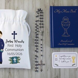 K&ouml;nnte beinhalten: Ein Erstkommunion-Set mit einem wei&szlig;en Kordelzugbeutel mit einer Jungenillustration und dem Namen "John Woods". Enth&auml;lt einen Rosenkranz, ein blaues "My Mass Book" und eine Gl&uuml;ckwunschkarte. Das Datum "16/05/2026" ist auf der Tasche aufgedruckt.