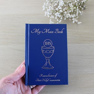K&ouml;nnte beinhalten: Ein blaues "My Mass Book" mit goldfarbenen Buchstaben und einer Kelchillustration. Das Buchcover enth&auml;lt auch den Text "Remembrance of First Holy Communion". Das Buch wird in einer Hand gehalten.