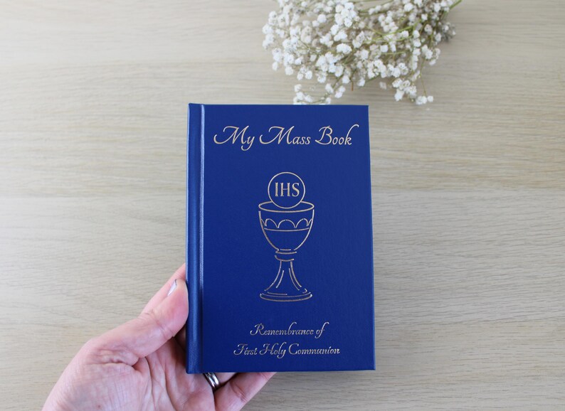 K&ouml;nnte beinhalten: Ein blaues "My Mass Book" mit goldfarbenen Buchstaben und einer Kelchillustration. Das Buchcover enth&auml;lt auch den Text "Remembrance of First Holy Communion". Das Buch wird in einer Hand gehalten.