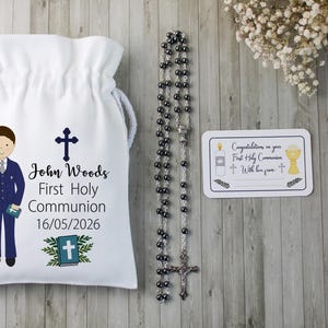 K&ouml;nnte beinhalten: Ein wei&szlig;er Kordelzugbeutel mit einer Cartoon-Illustration eines Jungen in einem blauen Anzug, einem Kreuz und dem Text "John Woods Erste Heilige Kommunion 16/05/2026". Ein Rosenkranz und eine Karte mit religi&ouml;sen Symbolen sind ebenfalls vorhanden.
