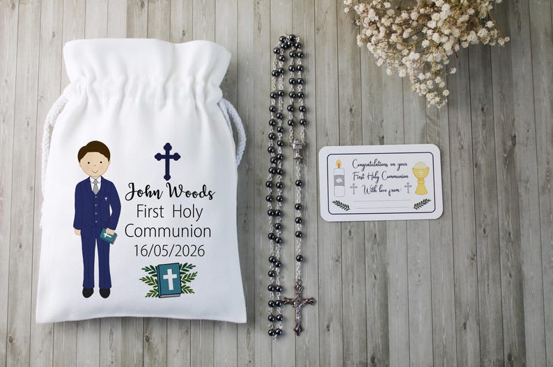 K&ouml;nnte beinhalten: Ein wei&szlig;er Kordelzugbeutel mit einer Cartoon-Illustration eines Jungen in einem blauen Anzug, einem Kreuz und dem Text "John Woods Erste Heilige Kommunion 16/05/2026". Ein Rosenkranz und eine Karte mit religi&ouml;sen Symbolen sind ebenfalls vorhanden.