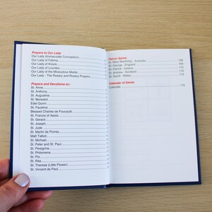 K&ouml;nnte beinhalten: Ein offenes Buch mit blauem Einband, das eine Liste von Gebeten und Andachten zeigt. Der Text enth&auml;lt "Gebete zu Unserer Lieben Frau" und "Gebete und Andachten zu". Das Buch wird von einer Hand gehalten.