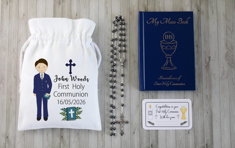 K&ouml;nnte beinhalten: Ein Erstkommunion-Set mit einem wei&szlig;en Kordelzugbeutel mit einer Jungenillustration und Text, einem Rosenkranz, einem blauen Buch mit dem Titel "My Mass Book" und einer Karte. Der Beutel tr&auml;gt den Namen "John Woods" und das Datum 16.05.2026.