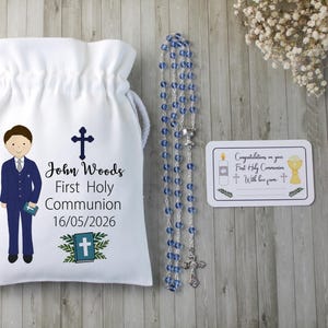 K&ouml;nnte beinhalten: Ein wei&szlig;er Kordelzugbeutel mit einer Cartoon-Illustration eines Jungen im Anzug, einem Kreuz und dem Text "John Woods Erste Heilige Kommunion 16/05/2026". Ein Rosenkranz mit blauen Perlen und eine Karte mit religi&ouml;sen Symbolen sind ebenfalls vorhanden.