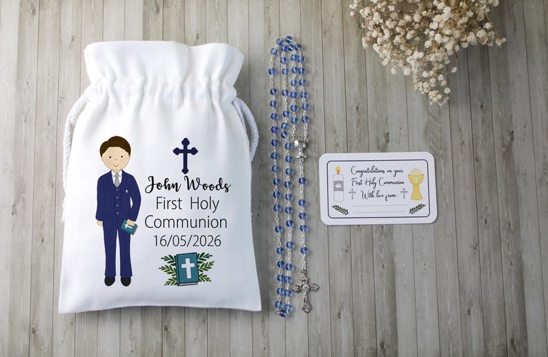 K&ouml;nnte beinhalten: Ein wei&szlig;er Kordelzugbeutel mit einer Cartoon-Illustration eines Jungen im Anzug, einem Kreuz und dem Text "John Woods Erste Heilige Kommunion 16/05/2026". Ein Rosenkranz mit blauen Perlen und eine Karte mit religi&ouml;sen Symbolen sind ebenfalls vorhanden.
