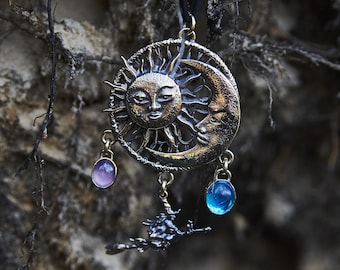 Puede incluir: Un collar con un colgante de plata con un diseño de sol y luna. El colgante tiene tres abalorios colgantes: una gema morada, una gema azul y un pequeño árbol.