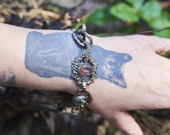 May include: A silver bracelet with a large, oval-shaped, red and clear gemstone. The bracelet is on a person's wrist with a black cat tattoo.