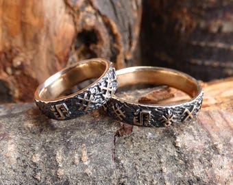 May include: Two silver rings with a textured surface and gold runes engraved on the band. The rings are on a wooden background.