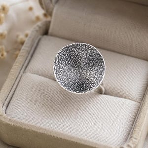 Op de afbeelding: Een zilveren ring met een grote, ronde en holle kop, voorzien van een donkere, complexe textuur met kriskras lijnen. De ring wordt gepresenteerd in een beige, met stof bekleed juwelendoosje met een scharnierend deksel.