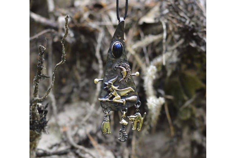 Close-up view of a mixed metal witch pendant with a blue lapis lazuli cabochon, detailed witch on broomstick, crescent moon, and cat familiar charms, showcasing intricate craftsmanship.