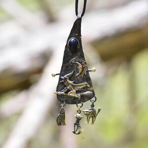Detailed view of a Wiccan-inspired necklace pendant featuring a witch riding a broomstick, crescent moon, and three dangling charms representing cat familiars and a cauldron, set against a blurred natural background.