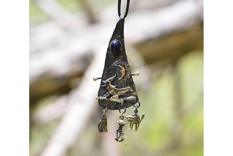 Detailed view of a Wiccan-inspired necklace pendant featuring a witch riding a broomstick, crescent moon, and three dangling charms representing cat familiars and a cauldron, set against a blurred natural background.