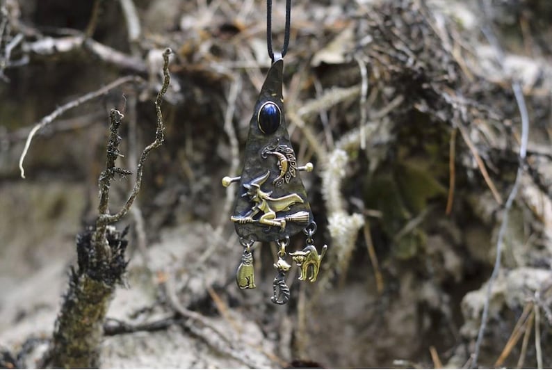 Handcrafted witch pendant necklace featuring a blue lapis lazuli gemstone, crescent moon, and witch on broomstick design with dangling cat and cauldron charms, set against a natural forest background.