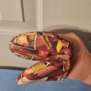 May include: A pair of oven mitts with a vintage postage stamp design. The mitts feature a repeating pattern of stamps in shades of red, brown, and beige, with the text "USA" visible. The text "BE SURE TO DO" is also visible.