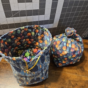 Puede incluir: Dos bolsas de dados azul marino, una abierta y otra cerrada, sobre una superficie de madera. Las bolsas presentan un patrón repetido de dados coloridos. La bolsa abierta está llena de varios dados, y el cordón es amarillo.