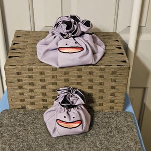 May include: Two light purple drawstring pouches featuring an embroidered face with two small black circular eyes and a wide, curved mouth with a pinkish-orange interior. The bags have a gathered top closure and a black interior lining.