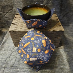 May include: Two navy blue bowl cozies with a bread-themed pattern. One cozy holds a dark bowl with a green interior. The cozies feature various bread illustrations and phrases like "I knead you" and "You crust me."